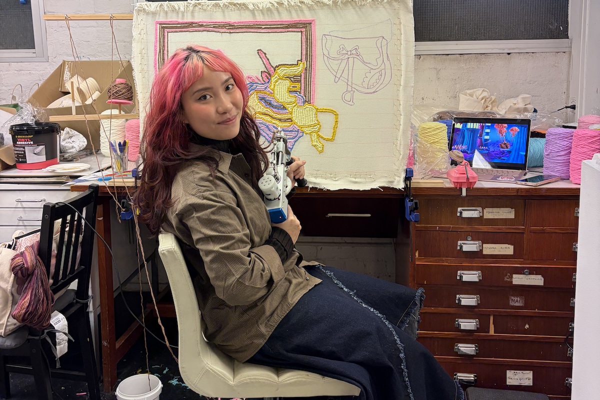 Artist Naomi Tang with pink-highlighted hair sitting in her studio holding a tufting gun, in front of an in-progress tufted textile artwork.